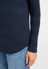 Granatowy sweter z dzianiny z teksturowanymi poziomymi paskami, długimi rękawami i zaokrąglonym dołem. Połączony z jasnoniebieskimi dżinsami.