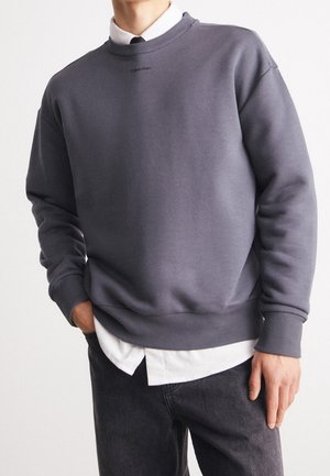 Sweatshirt gris avec des poignets et un ourlet côtelés, featuring un col rond et un petit logo sur la poitrine. Superposé sur une chemise blanche.