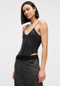 Γυναίκα με ίσια μαύρα μαλλιά που φοράει μαύρο αμάνικο τοπ με κουμπιά και σκούρα γκρι φούστα με μαύρη ζώνη, στέκεται μπροστά σε απλό φόντο.