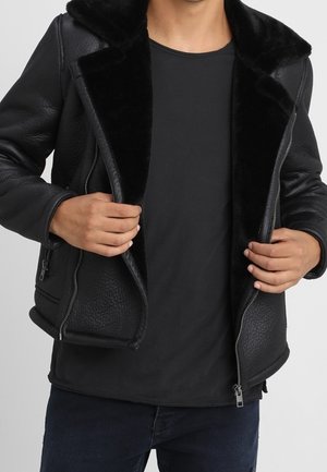 Mand iført sort fåreskindjakke med pelsforing, glidende lynlåsen ned over en simpel sort skjorte og mørke jeans.