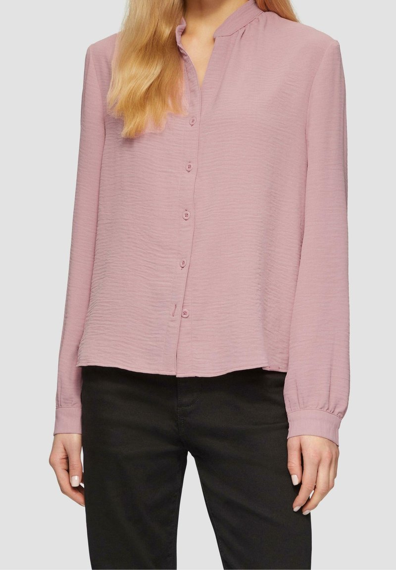 Femme portant un chemisier à manches longues rose clair avec un col mao et un pantalon noir, posant devant un fond uni.