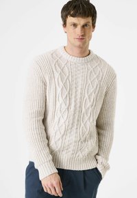 Mann trägt cremefarbenen grob gestrickten Pullover und dunkle Hose, steht mit einer Hand in der Tasche vor weißem Hintergrund.