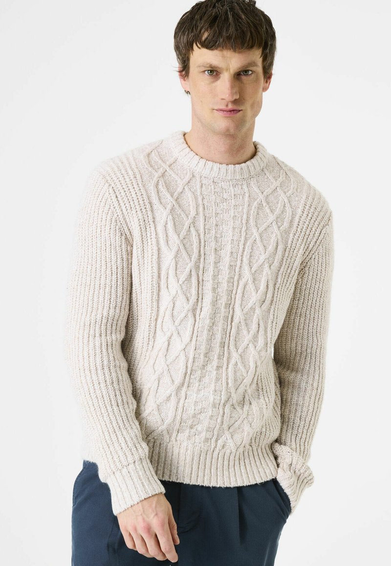 Mann trägt cremefarbenen grob gestrickten Pullover und dunkle Hose, steht mit einer Hand in der Tasche vor weißem Hintergrund.