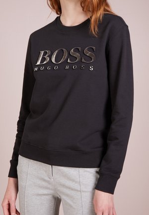 Kobieta ubrana w czarną bluzę z dużym logo "BOSS HUGO BOSS" i jasnoszare spodnie na neutralnym tle.