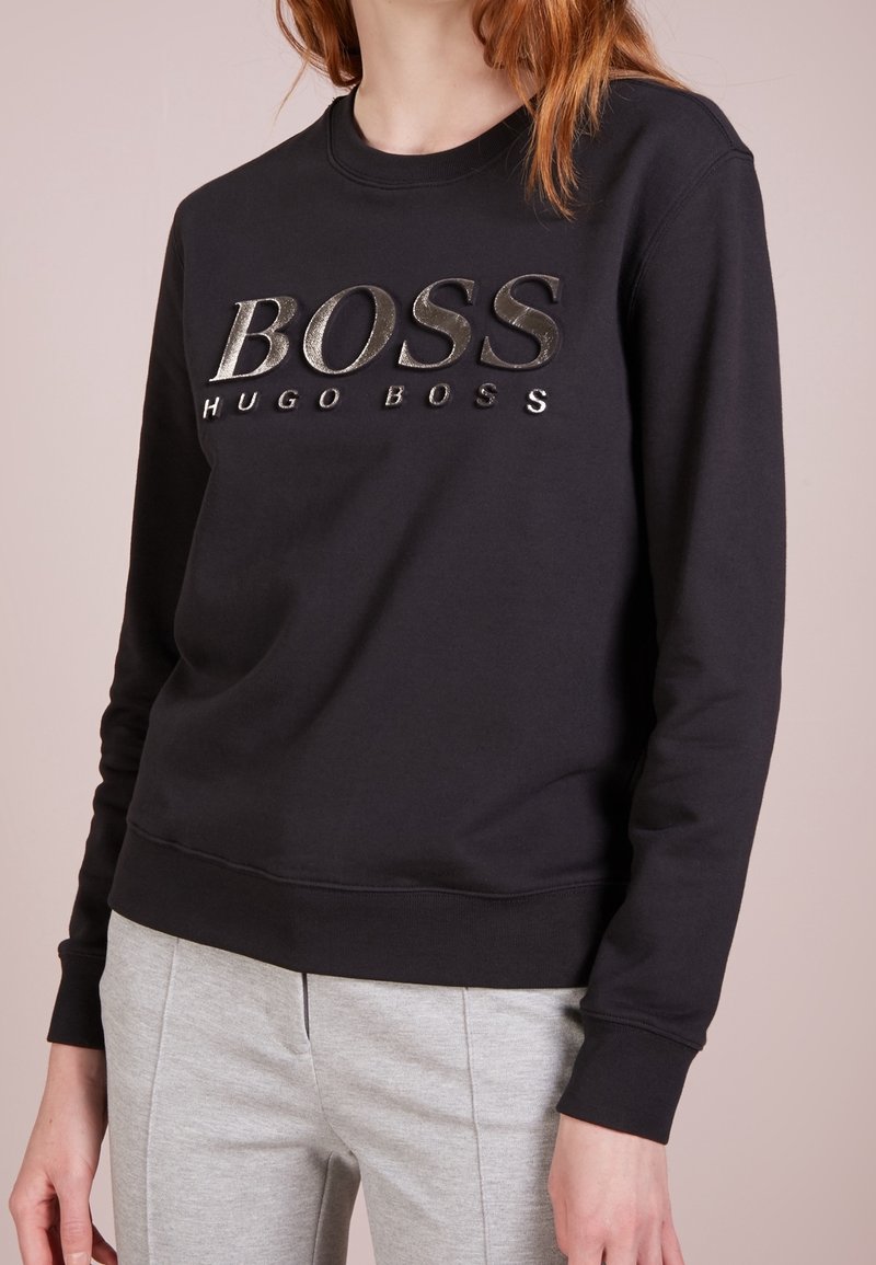 Femme portant un sweat-shirt noir avec un grand logo "BOSS HUGO BOSS" et un pantalon gris clair sur fond neutre.