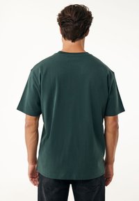 Tmavě zelené bavlněné tričko s volným střihem, krátkými rukávy, kulatým výstřihem a hladkou, jednotnou texturou. Jednoduchý design na zadní straně.