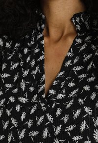 Haut noir avec un col en V, un col froncé et des motifs de feuilles blanches sur un tissu texturé avec de subtiles lignes verticales.