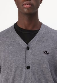 Cardigan gri cu design în formă de V, două nasturi negri și un mic logo pe partea stângă. Textură netedă, vedere de aproape a părții superioare a corpului.