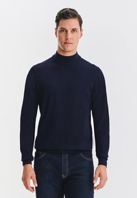 Granatowy sweter z półgolfem wykonany z miękkiego, gładkiego dziania. Posiada długie rękawy oraz ściągacze na mankietach i u dołu. Noszony z ciemnymi jeansami.