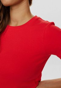 Haut en rouge à côtes avec un col rond et des manches longues. Le tissu est lisse avec une silhouette ajustée, mettant en valeur des détails texturés.