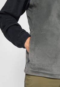 Szürke testű, fekete ujjú fleece mellény. Simább, puha textúrával, elasztikus mandzsettákkal és oldalsó cipzáros zsebbel rendelkezik.