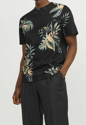 Homme portant une chemise noire à manches courtes avec des imprimés de feuilles tropicales beige et vertes et un pantalon noir, debout avec une main dans la poche.
