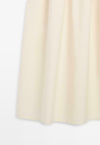 Partie inférieure d'une jupe couleur crème avec des plis doux et une texture de tissu lisse sur un fond blanc uni.