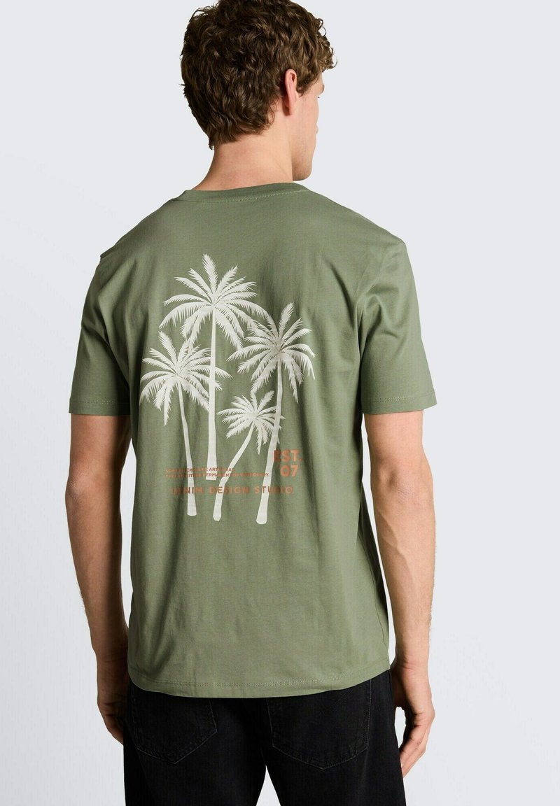 Mann trägt olivgrünes T-Shirt mit weißem Palmenbaum-Grafik und Text auf dem Rücken, kombiniert mit schwarzen Hosen, mit dem Rücken zur Kamera.