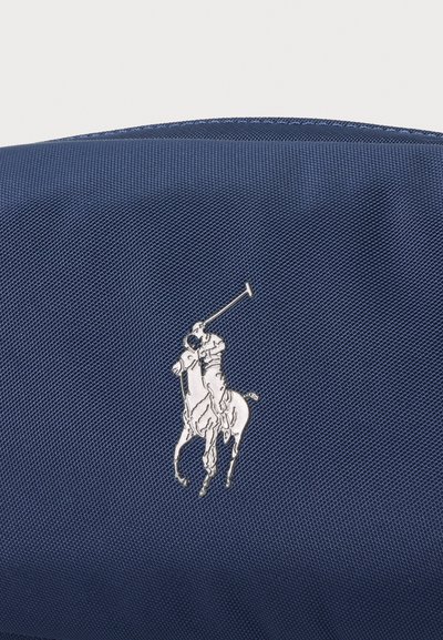 Ασημένιο λογότυπο παίκτη πόλο σε άλογο που κρατάει ανασηκωμένο μαλέτο σε υφασματένιο σκουρό μπλε φόντο με υφή.