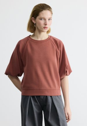 Γυναίκα με κοντά ξανθά μαλλιά, φοράει τοπ με μανίκια-φουσκωτά σε σκουρόχρωμο πορτοκαλί και άνετο γκρι παντελόνι, στέκεται μπροστά σε απλό λευκό φόντο.