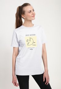 Bílé bavlněné tričko s černým liniov�ým obrázkem holubice na žlutém pozadí, s nápisem „PICASSO“ nahoře a textem dole.