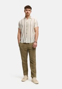 Camisa de manga corta y cuello con rayas verticales en beige y marrón. Combinada con pantalones texturizados en verde oliva y zapatillas de color claro.
