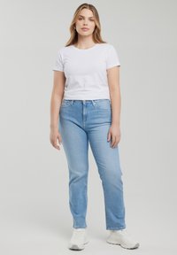 Jauna sieviete stāv, tērpusies vienkāršā baltā īsroku krekliņā, gaisā zilos džinsu biksēs un baltos sporta apavos, pret vienkāršu gaiši pelēku fonu.