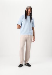 Lys blå teksturert kortermet skjorte kombinert med beige ribbede bukser og svarte sneakers med hvite striper; stående mot en hvit bakgrunn.