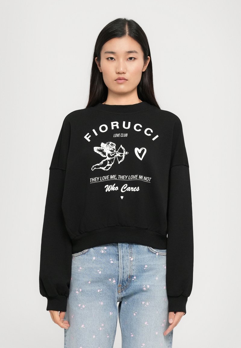 Jovem mulher vestindo um moletom preto com um cupido, coração e o texto "Fiorucci Love Club, Eles me amam, eles não me amam, Quem se importa" e jeans de tom azul claro.