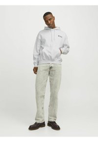 Un sweat à capuche gris clair avec une poche avant et un cordon de serrage, associé à un jean clair délavé et des bottes foncées, se tenant sur un fond neutre.
