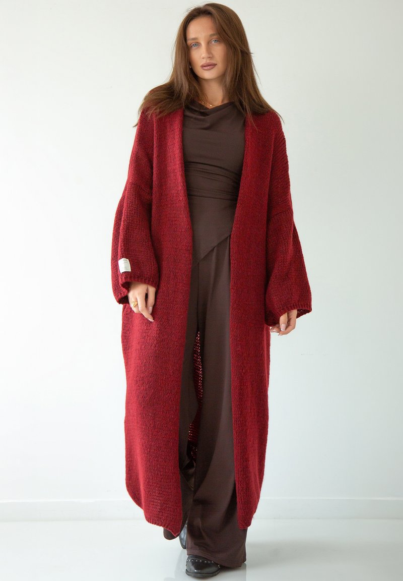 Czerwony sweter dzianinowy z szerokimi rękawami i otwartym przodem, nałożony na brązowy element odzieży. Tekstura wygląda na przytulną i miękką.