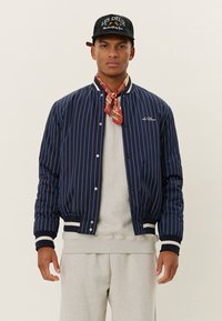 Navy-blue striped bomber jacket with ribbed cuffs and hem, white accents, worn over a light gray sweater and complemented by a patterned scarf.