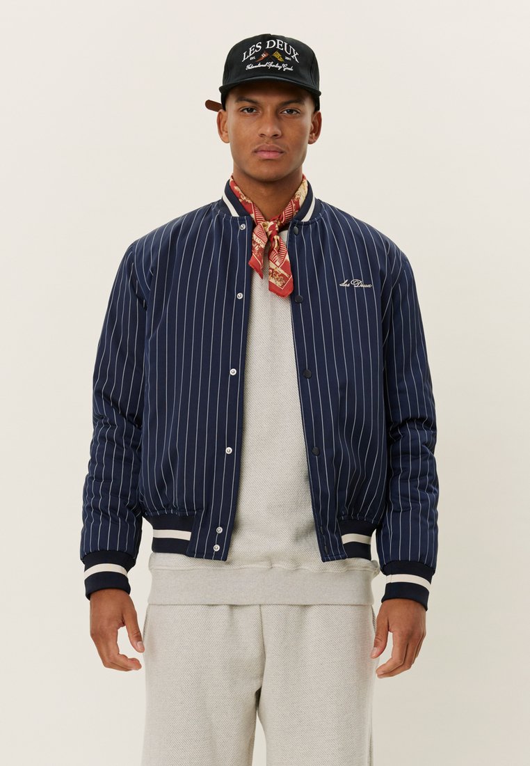 Navy-blue striped bomber jacket with ribbed cuffs and hem, white accents, worn over a light gray sweater and complemented by a patterned scarf.