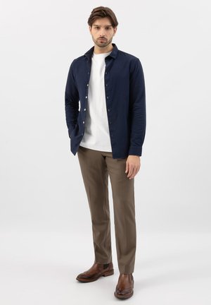 Homme debout portant une chemise bleu marine à boutons sur un t-shirt blanc, un pantalon marron et des chaussures en cuir marron sur un fond blanc uni.