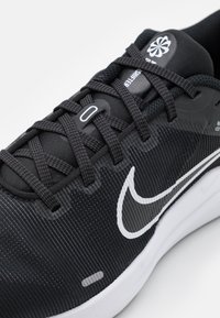 Crna atletska cipela s teksturiranim mrežastim gornjim dijelom, glatkim detaljima, istaknutim bijelim Nike logotipom i ravnim vezicama za sigurno prianjanje.