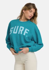 Turkusowy sweatshirt z napisem "SURF" w jasnoniebieskim kolorze, luźny krój, okrągły dekolt, ściągacze przy rękawach i miękka faktura, noszony z beżowymi spodniami z kordurowej tkaniny.