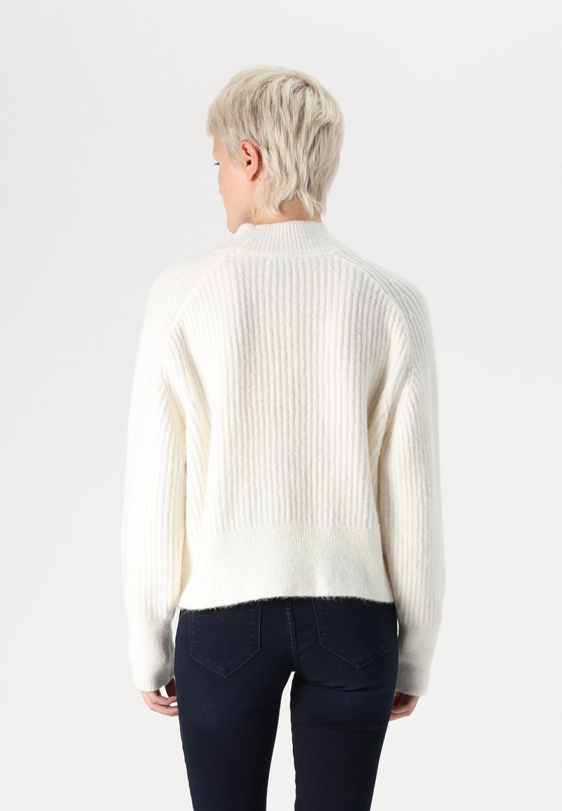 Maglione lavorato a maglia color crema con texture a coste, scollo rotondo e orlo leggermente accorciato, abbinato a jeans blu scuro.