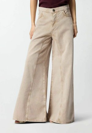 Pantaloni beige a gamba larga e vita alta con cuciture visibili, indossati da una persona con le mani rilassate ai lati, abbinati a sandali aperti e una maglietta color granata.