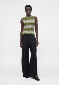 Γυναίκα στέκεται φορώντας αμάνικη πράσινη μπλούζα με σχέδιο, φαρδιά μαύρη παντελόνια και μαύρα loafers με χρυσή λεπτομέρεια σε απλό λευκό φόντο.