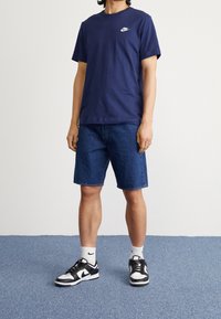 Marinblå bomulls T-shirt med en vit logotyp, tillsammans med denimshorts i knälängd och svart- och vitaskor med vita strumpor.