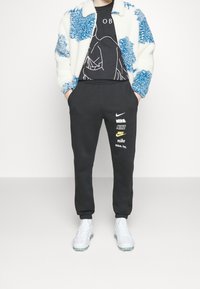 Chaqueta azul y blanca difusa sobre una camiseta gráfica negra; pantalones de chándal negros de Nike con logotipos; zapatos deportivos transparentes.