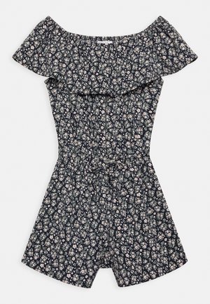 Kombinezon z odkrytymi ramionami, kwiatowym wzorem, falbanką przy dekolcie, taliowaniem oraz ciemnym tłem ozdobionym drobnymi białymi i jasno różowymi kwiatami.