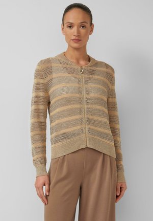 Frau trägt eine Strickjacke mit Reißverschluss in Beige und Gold mit Metallstreifen, langen Ärmeln, und eine hochtaillierte hellbraune Hose.