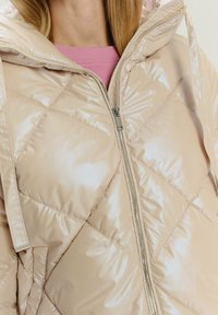 Giacca a vento beige imbottita con texture lucida e trapuntata, cappuccio e zip argento. Indossata sopra un indumento rosa.