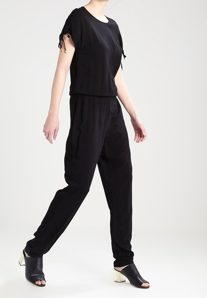 Combinaison noire à manches courtes, avec une taille élastique et des détails de cordon de serrage. Le modèle porte des chaussures noires ouvertes à l'avant avec un talon métallique.