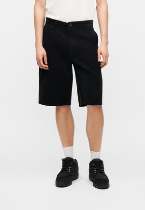 Person iført sorte knælange shorts, hvide sokker, sorte snøresko og en hvid indstukket skjorte, stående mod en ensfarvet baggrund.