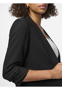 Blazer noir au design élégant, avec des manches trois-quarts, un col châle et une texture lisse. Le haut blanc en dessous est partiellement visible.