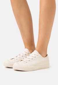 Plátěné tenisky v krémové barvě, s kulatou špičkou a žebrovanou gumovou podrážkou. Obsahují tradiční design s šněrováním a kovové očka na tkaničky.