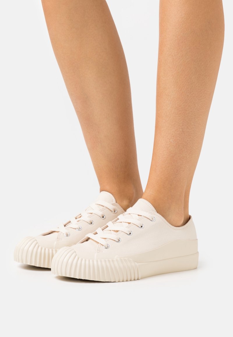 Plátěné tenisky v krémové barvě, s kulatou špičkou a žebrovanou gumovou podrážkou. Obsahují tradiční design s šněrováním a kovové očka na tkaničky.