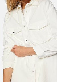 Biała koszula z korduroyu ma zapięcie na guziki z przodu, dwie kieszenie na piersi z zapięciem na zatrzaski oraz podwinięte rękawy ukazujące teksturowany materiał.