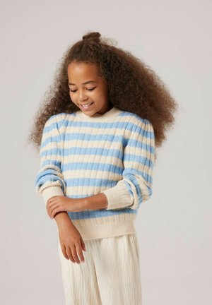 Fille aux cheveux bouclés volumineux portant un pull rayé bleu et crème et un pantalon crème texturé, souriant et regardant son bras.