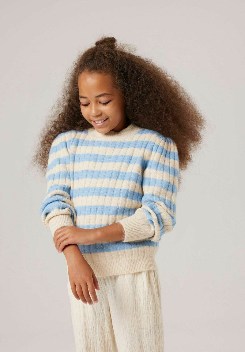Mädchen mit voluminösen, lockigen Haaren, das einen blau- und cremefarben gestreiften Pullover und cremefarbene, strukturierte Hose trägt. Sie lächelt und schaut auf ihren Arm.