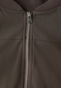 Brun jacka med metalldragkedja, ribbad krage och slät tyg med subtila sömnadsdetaljer. Närbild av framsidan.