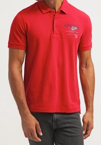 Homme portant un polo rouge à manches courtes avec un petit motif et un texte sur le côté gauche de la poitrine, assorti à un jean foncé.
